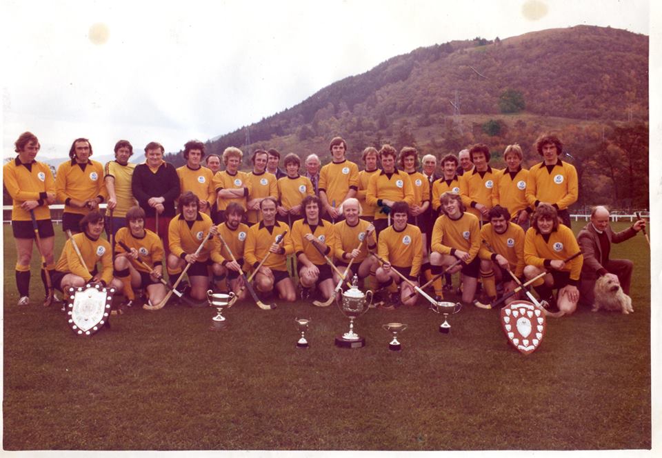 Colour image of Fort William's 1976-77 team in a yellow strip. Line up with the trophies they won that year