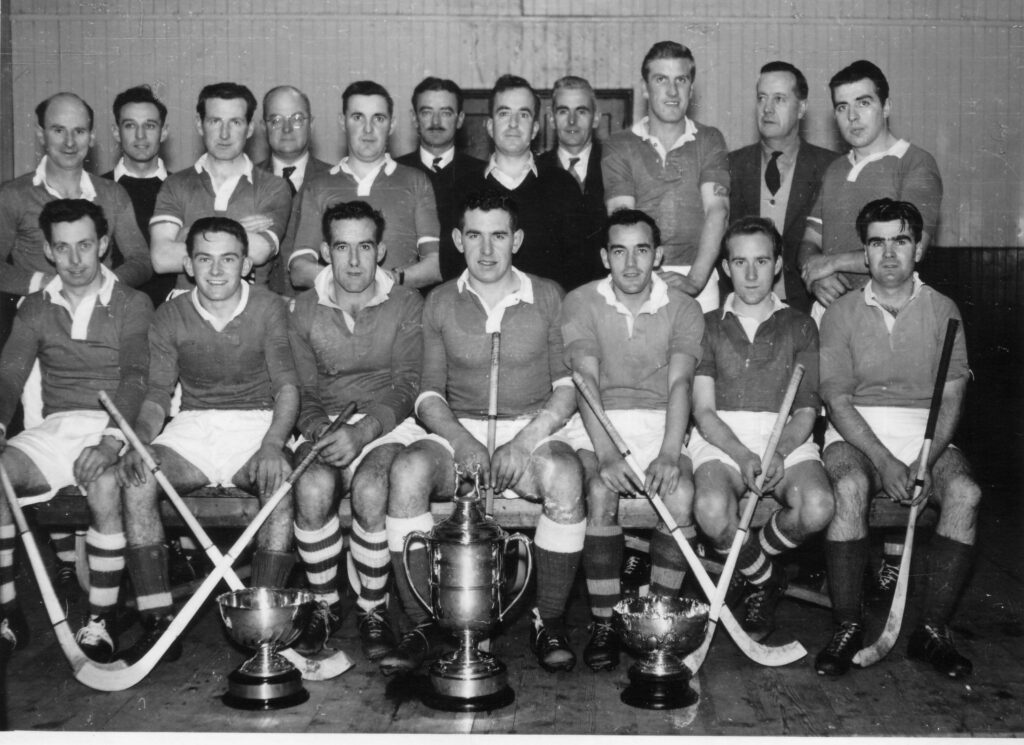 Black and white image of Kilmallie Shinty Team lined up with their shinty sticks and the Camanachd Cup