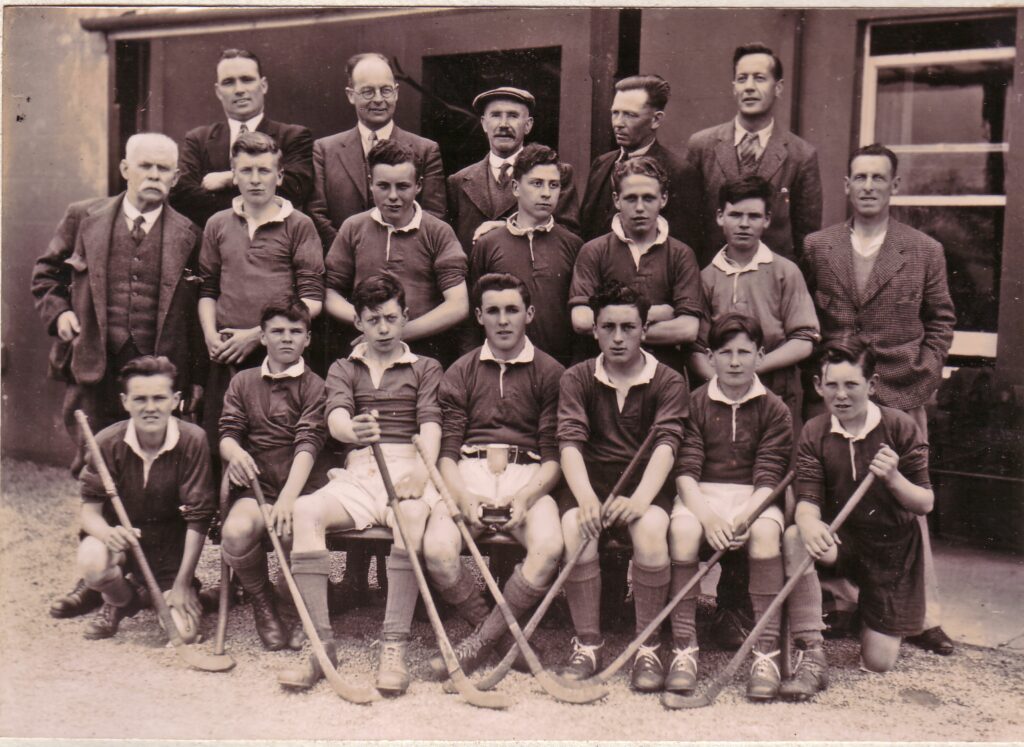 Black and white photograph of the youth team of 1947. The players are sat and standing for a team photograph with their shinty sticks and the trophy held by the captain in the centre of the phtograph