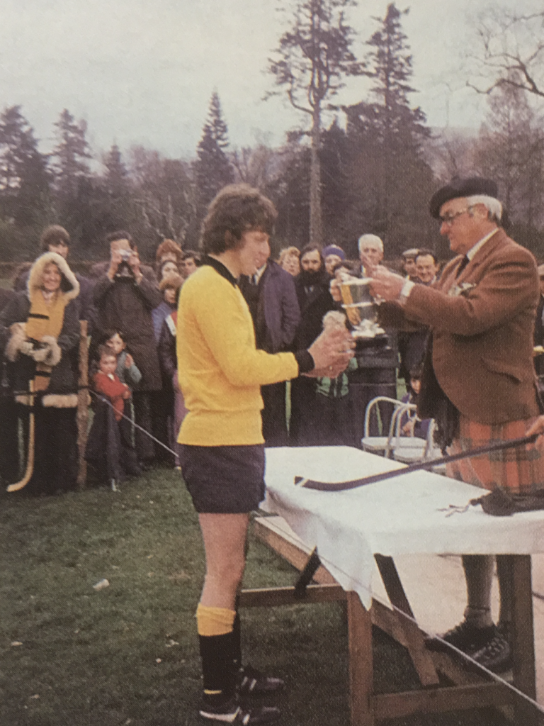 Colour image of Fort William player, Jo Smith, in his yellow strip collecting the Sutherland Cup trophy from Walter Cameron