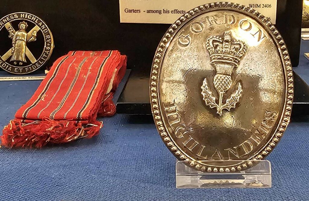 Inset of a museum case with red cloth garters to the left and a silver military cross belt plate to the right