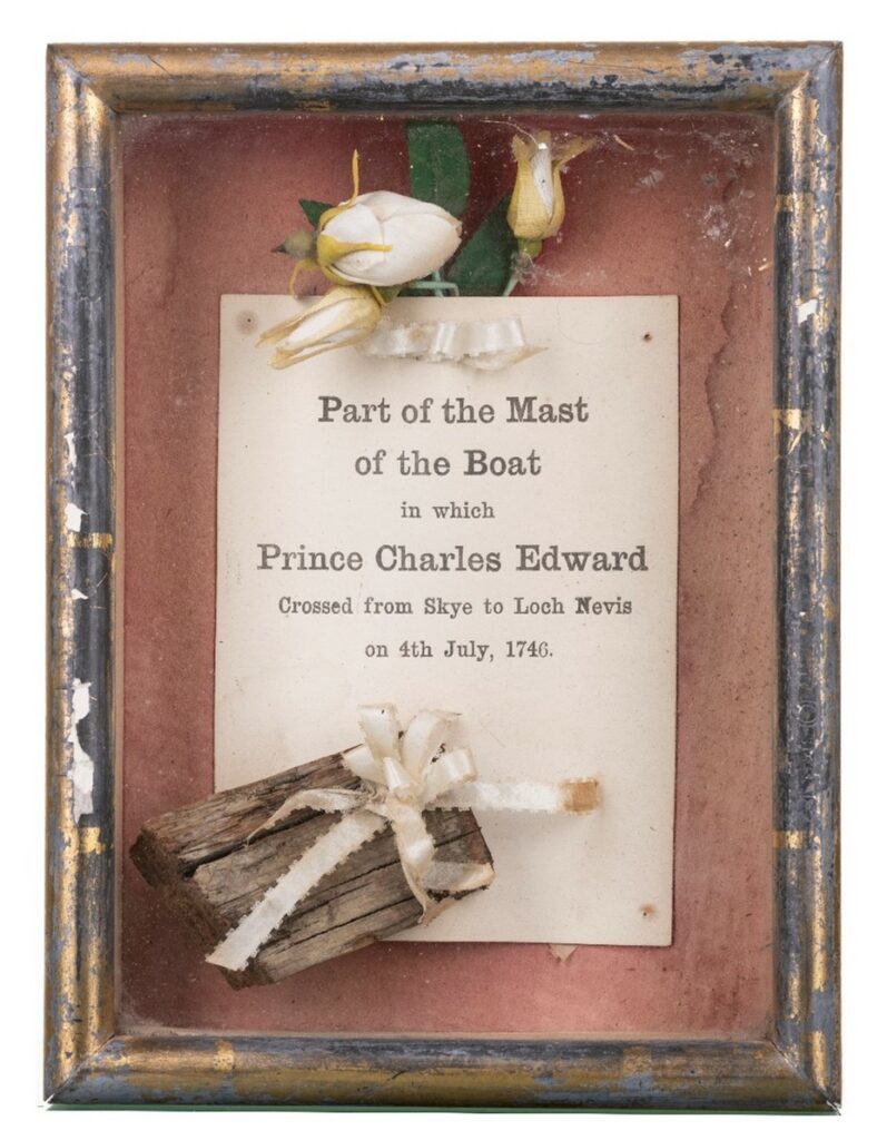 a small wooden frame containing a written statement which reads "Part of the mast of the boat in which Prince Charles Edward crossed from Skye to Loch Nevis on 4th July 1746" at the bottom of the freame is a small piece of weathered wood tied with a ribbon. At the top of the frame are white flowers