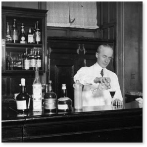 Black and white photograph of a man mixing a cocktail behind a bar. A number of bottles line the bar