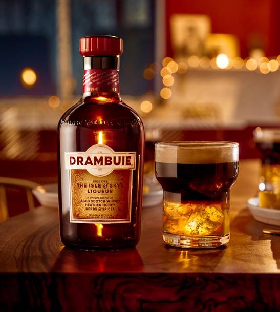 A bottle of Drabuie as it is today with its red top and gold label. To the right side of the bottle sits a glass of Drambuie as a cocktail on ice